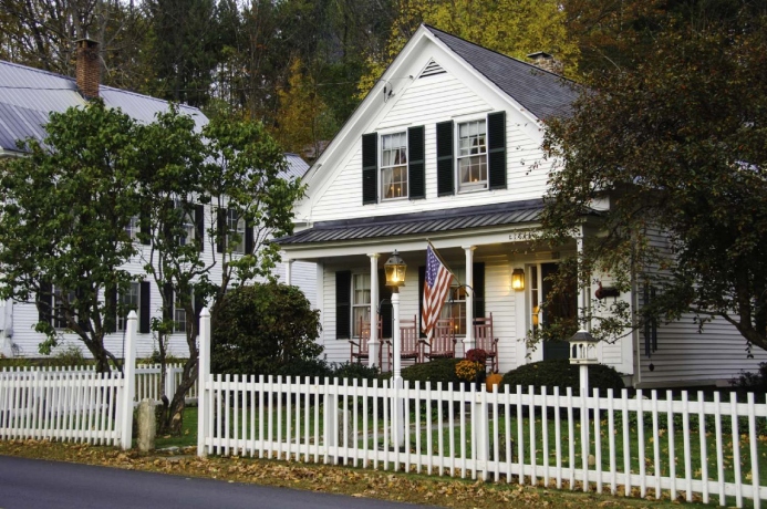 Charming white house with black shutters, American flag, and a picket fence, surrounded by trees and autumn foliage.